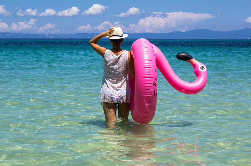 Lading standing in ocean with flamingo float on her shoulder and holding her hat