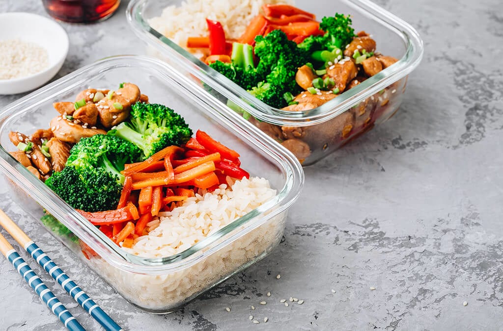 Meal prepared in glass containers with sesame chicken, broccoli, carrots and rice with chopsticks laying besides them